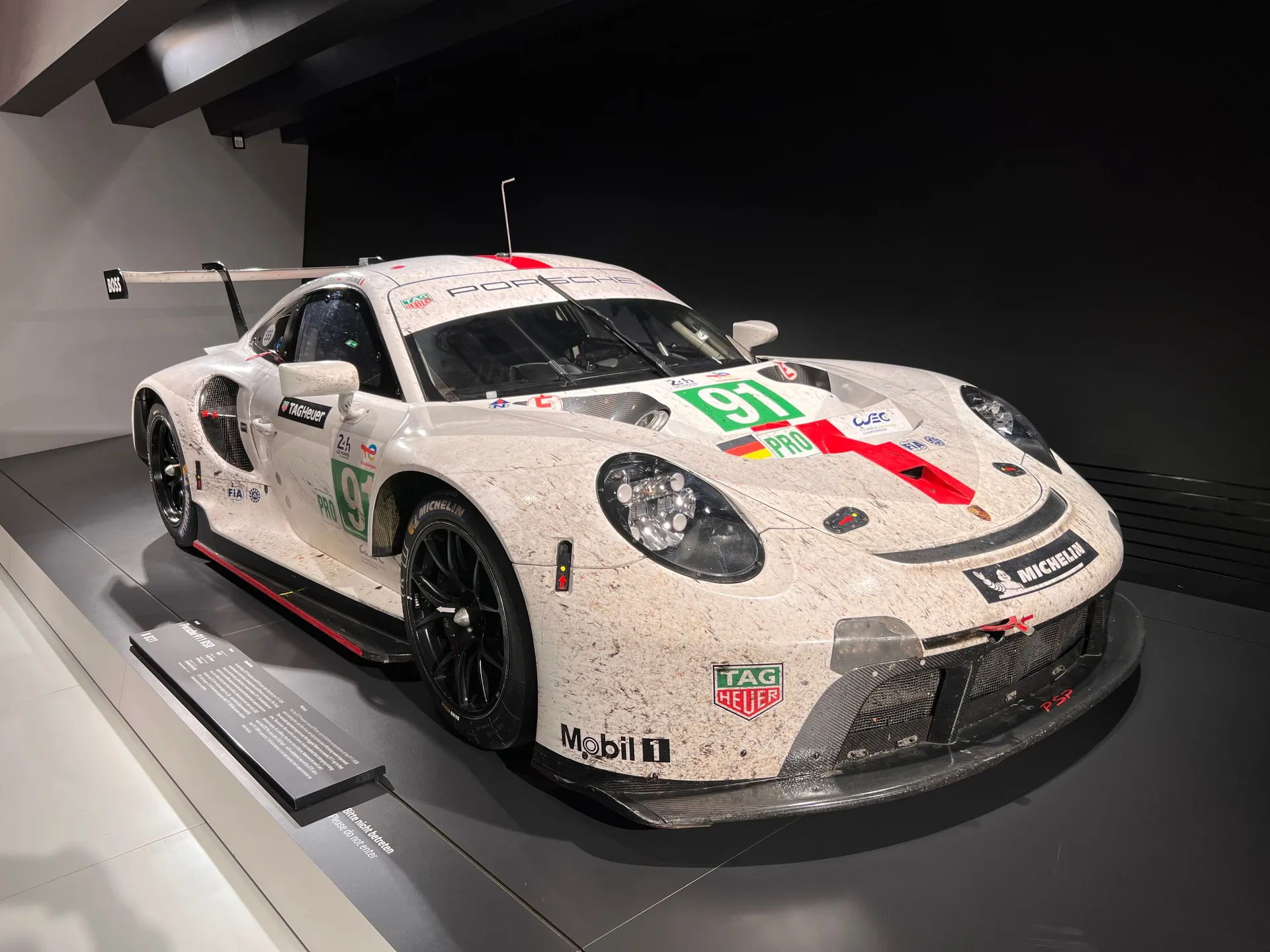 A raced Porsche 911 RSR, number 91, on a museum plinth with the dirt of the race still on it.