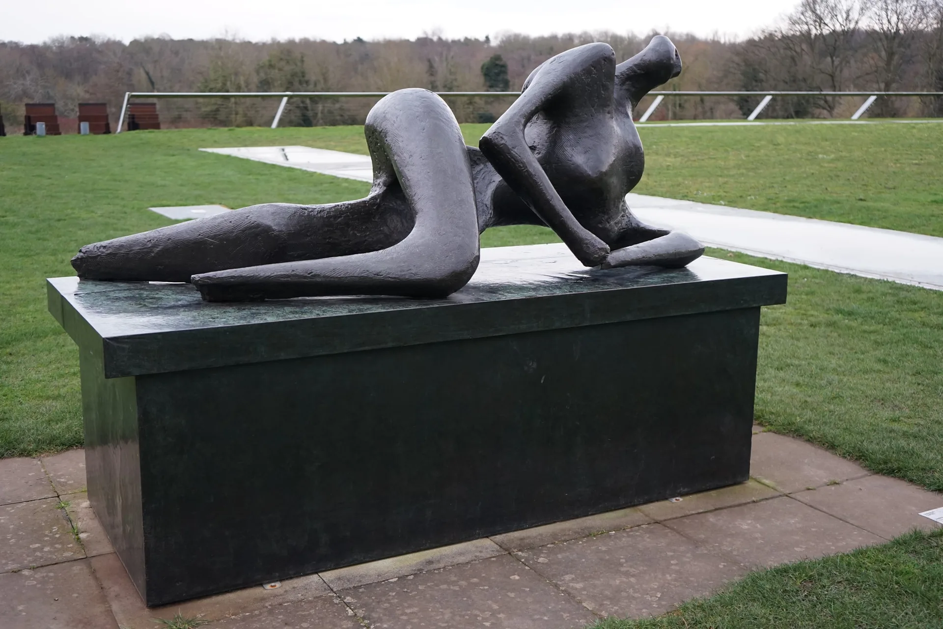 A reclining bronze figure on a stone plinth, set on a lawn under bare trees.